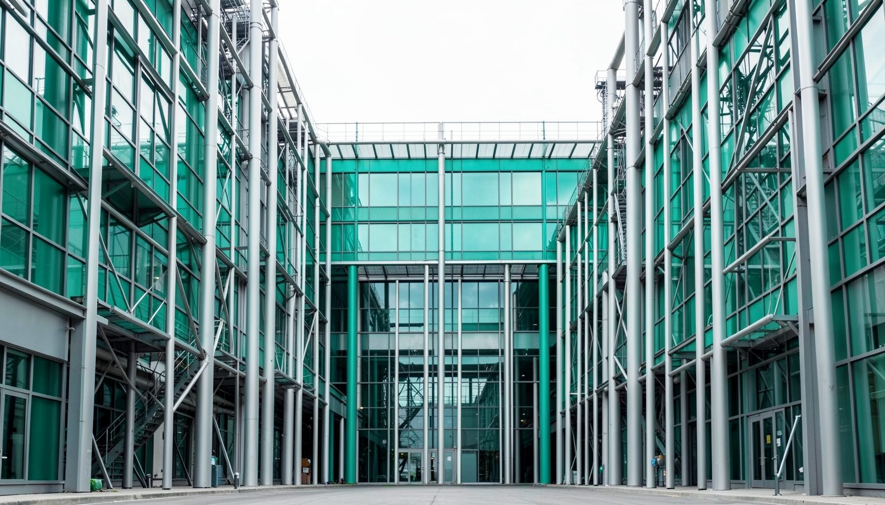 ภาพถ่ายสถาปัตยกรรมโรงงานอุตสาหกรรมแนวสมมาตร โทนสีเทาเขียวและเงิน ตัดกับเส้นสายของโครงสร้างเหล็กและกระจก